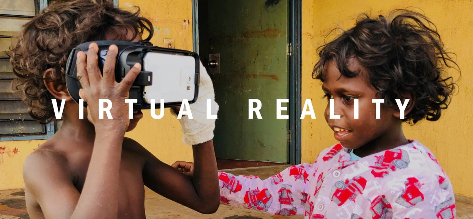 Virtual Reality: two children outside a yellow-walled building, one holding a VR headset to his eyes while the other watches and smiles