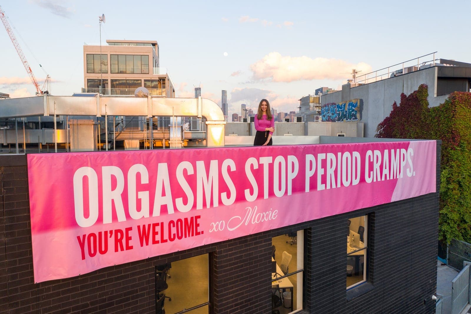 A woman stands on a city rooftop above a pink Moxie banner reading Orgasms stop period cramps. You're welcome. xo Moxie